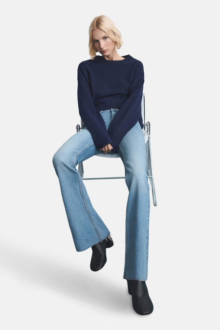 Vrouw zit op stoel, donkerblauwe trui, lichtblauwe jeans, zwarte laarsjes.