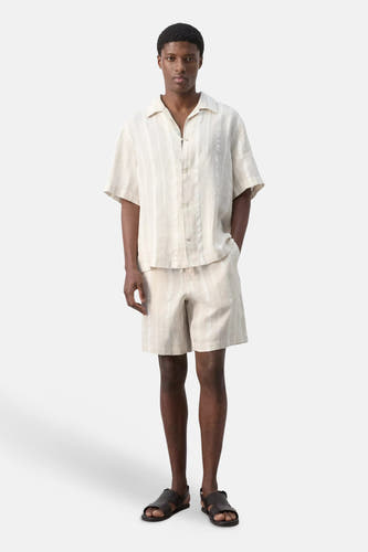 Chemise beige à manches courtes de MANGO à rayures verticales, portée par un mannequin avec un short rayé assorti et des sandales foncées.