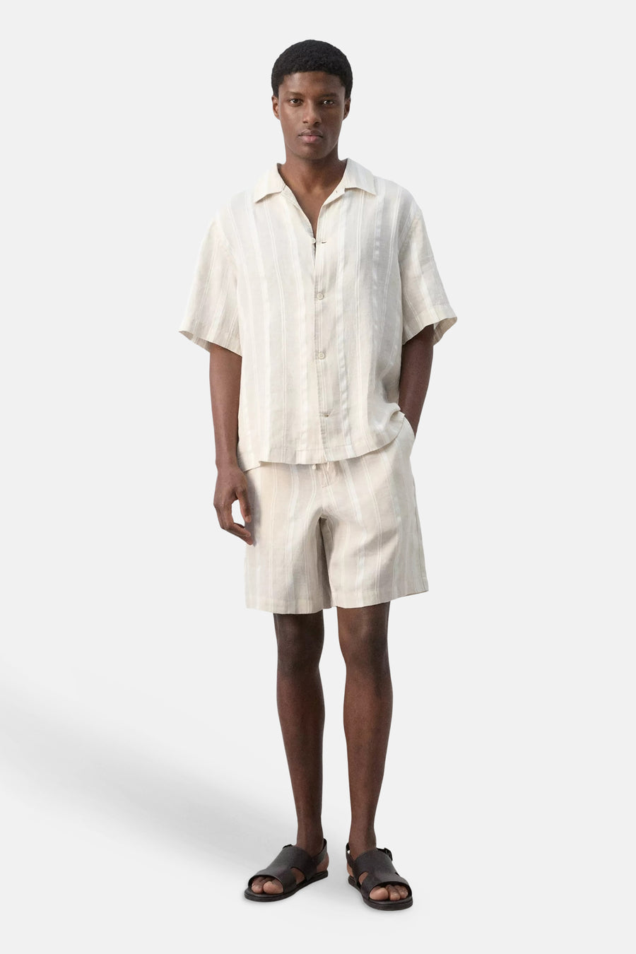 Chemise beige à manches courtes de MANGO à rayures verticales, portée par un mannequin avec un short rayé assorti et des sandales foncées.