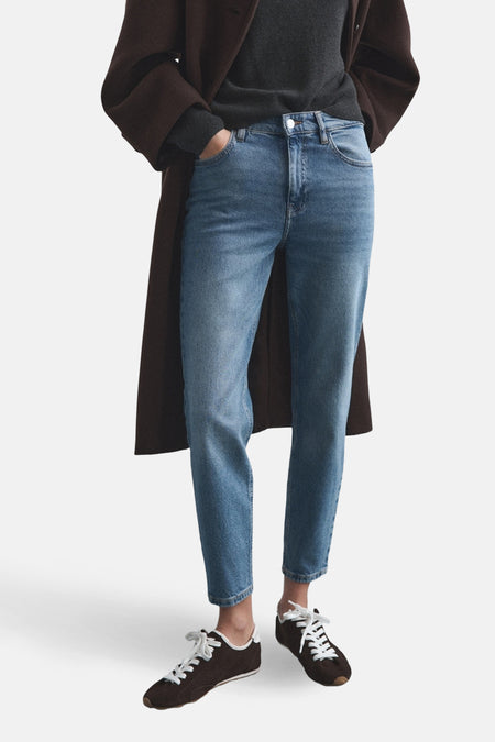 Jean mom en denim bleu de MANGO, avec des poches arrière et porté avec un pull gris et des chaussures marron foncé.