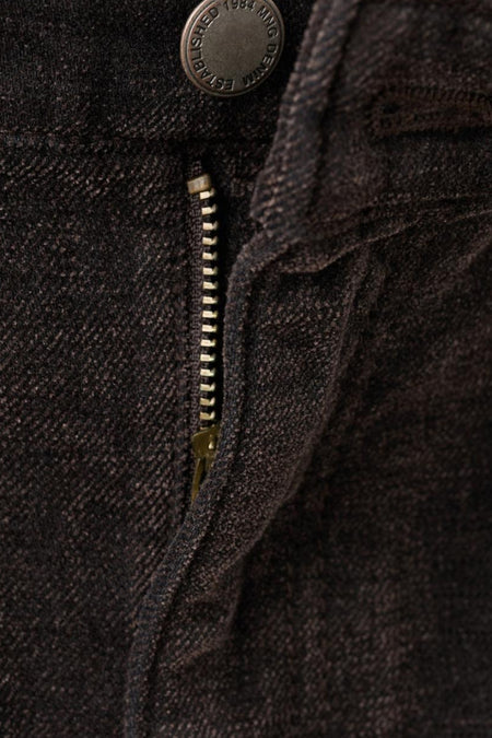 Detailopname van een donkerbruine jeans met een gedeeltelijk open rits en een knoop met "MNG DENIM" erop.