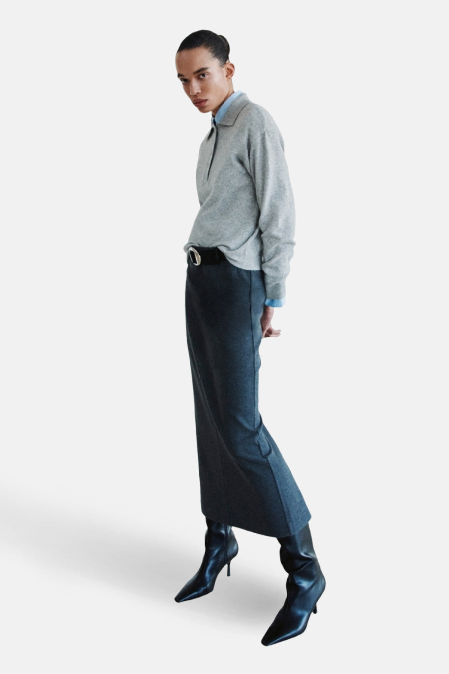Sweat-shirt gris à col rond de MANGO, porté avec une longue jupe, une ceinture et des bottes noires à talon.