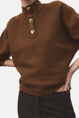 Pull marron avec col montant de MANGO, texture côtelée et manches bouffantes, associé à un pantalon à carreaux.