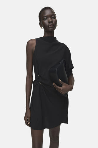 Robe noire sans manches de MANGO, avec une épaule dénudée, jupe courte et talons.