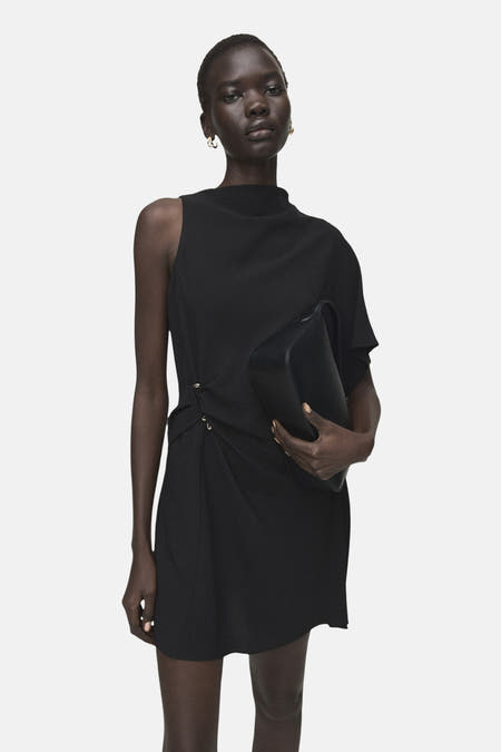 Robe noire sans manches de MANGO, avec une épaule dénudée, jupe courte et talons.