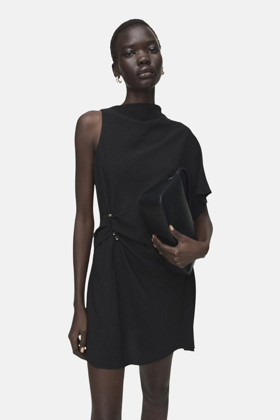 Robe noire sans manches de MANGO, avec une épaule dénudée, jupe courte et talons.