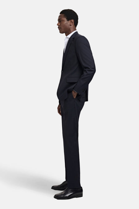 Pantalon de costume bleu MANGO, avec des poches et une texture lisse, en vue latérale, associé à un chemise blanche et des chaussures noires.