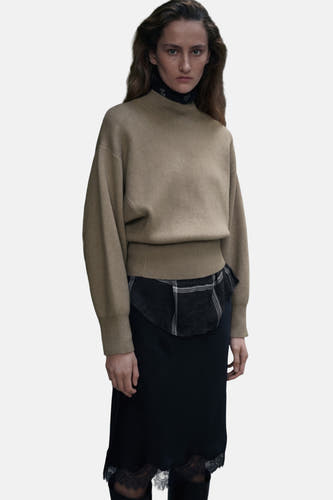Pull de kaki avec col rond de MANGO, associé à une jupe noire avec des détails froncés.