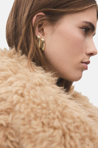 Boucles d'oreilles en or de MANGO, portées avec une veste douce et beige dans un profil latéral.