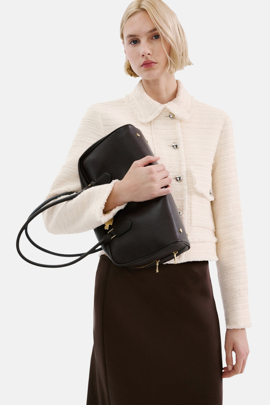 Vrouw in een crème jasje, bruine rok en zwarte handtas.