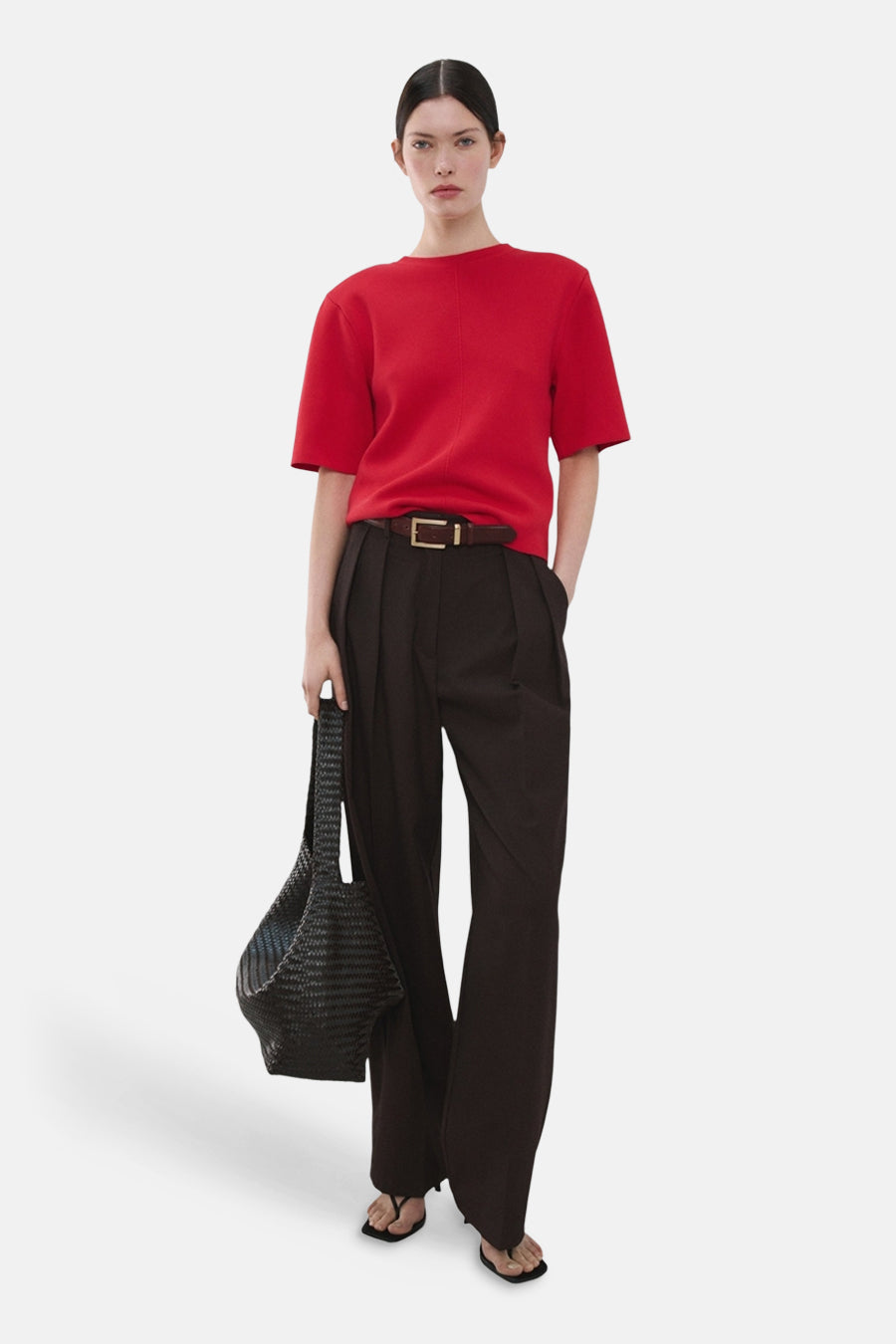 Pull rouge à col rond de MANGO, porté avec un pantalon marron, une ceinture et des sandales noires, avec un sac à main noir.