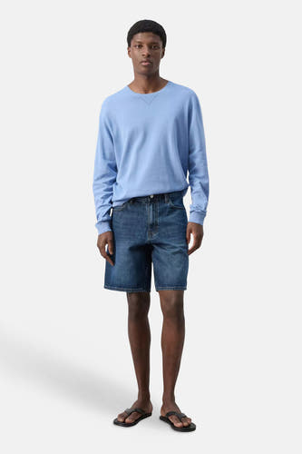 Bermuda en jean bleu marine de MANGO, porté par un homme debout vêtu d'un pull bleu clair et de sandales noires.
