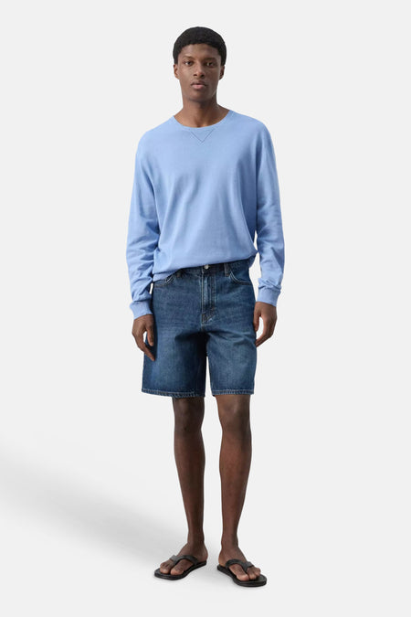 Bermuda en jean bleu marine de MANGO, porté par un homme debout vêtu d'un pull bleu clair et de sandales noires.