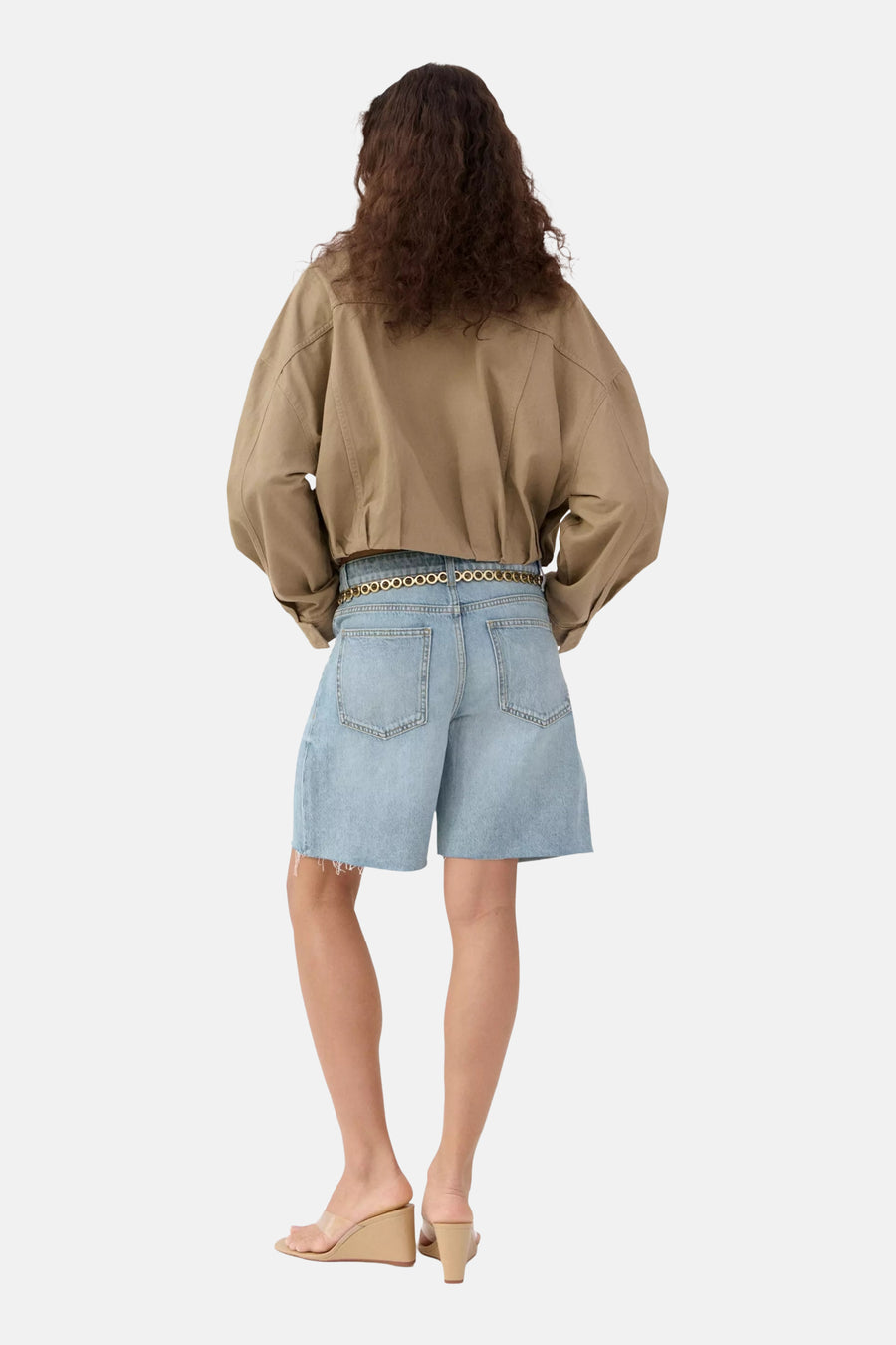 Lichtblauwe denim Bermuda van MANGO met onafgewerkte zoom, gecombineerd met een bruin overhemd en gouden kettingriem, van achteren gezien.