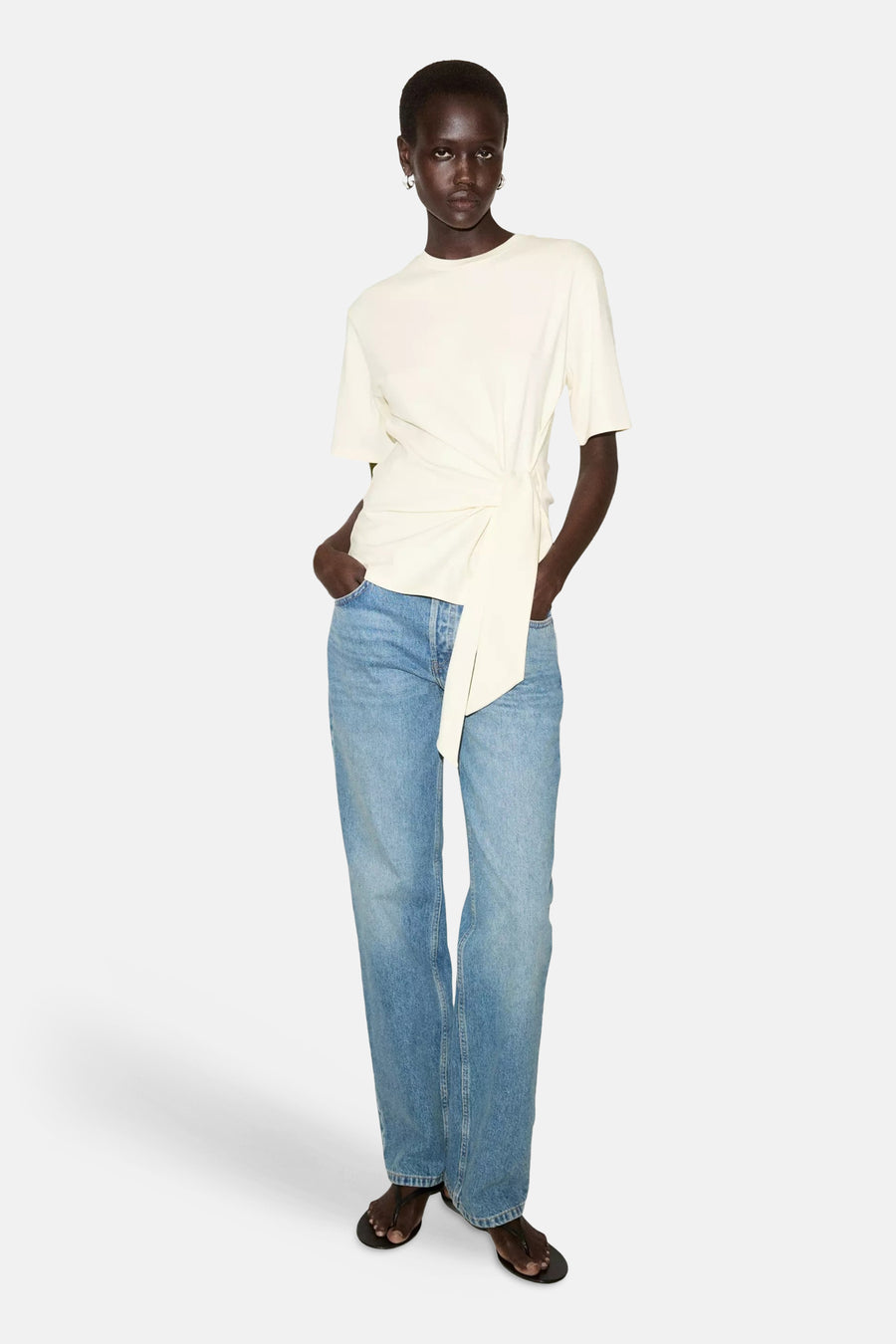 Gele korte mouwen t-shirt van MANGO met knoopdetail aan de zijkant, gedragen met blauwe jeans en zwarte sandalen, front view.