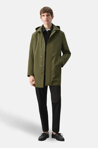 Long manteau kaki de MANGO, avec capuche, manches longues et porté ouvert sur une chemise noire et un pantalon.