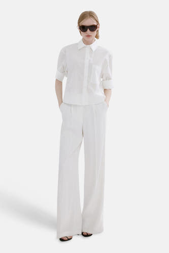 Chemise blanche à manches longues de MANGO, portée avec un pantalon blanc ample et des tongs noires.