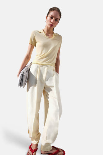 Polo jaune à manches courtes de MANGO avec col en V, porté par un mannequin avec un pantalon clair et des sandales rouges.
