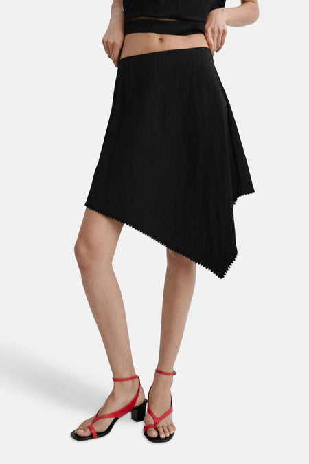 Jupe noire MANGO avec ourlet asymétrique et passepoil décoratif, portée avec une chemise blanche et des sandales marron.