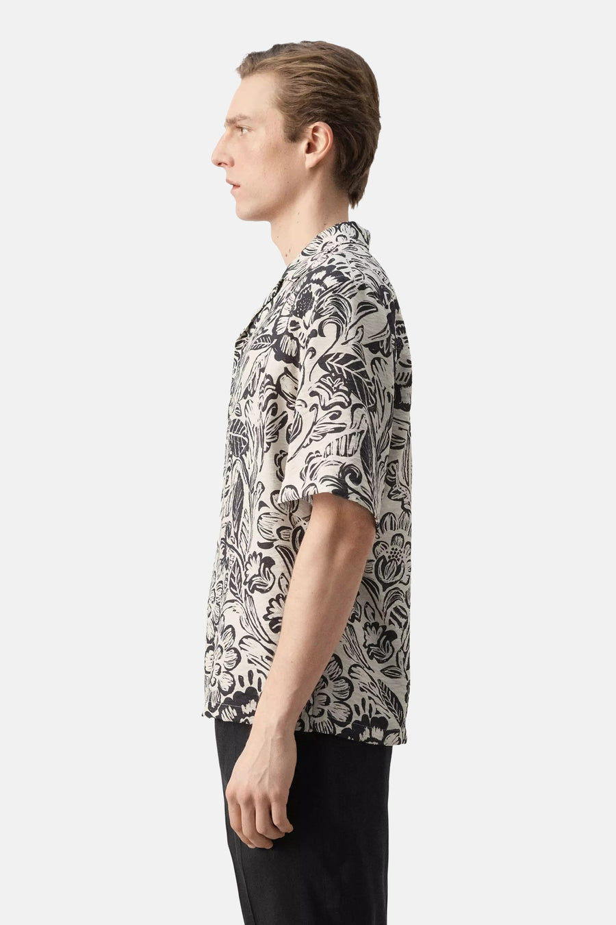 Chemise à manches courtes avec imprimé floral noir et blanc de MANGO, portée par un homme de profil avec un pantalon noir.