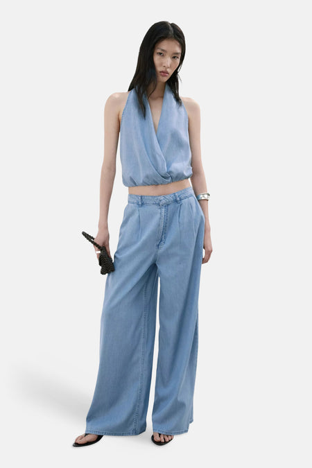 Lichtblauwe denim blouse zonder mouwen van MANGO, wrap-stijl met diep V-hals, gekenmerkt door een losse pasvorm en subtiele textuur, getoond in lifestyle-opstelling met bijpassende jeans en zwarte sandalen.