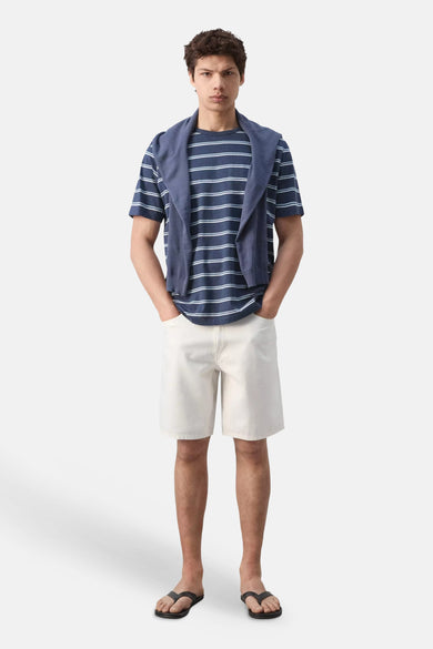 T-shirt bleu rayé de MANGO, porté avec un pull sur les épaules, un short blanc et des tongs.