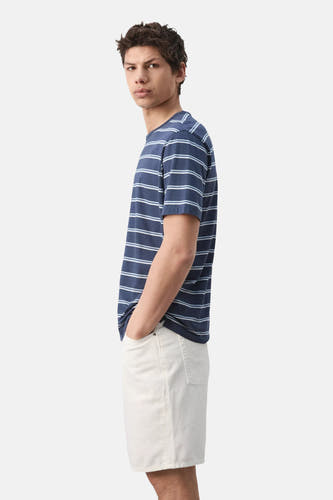 T-shirt rayé bleu de MANGO, avec des rayures blanches et bleu clair, manches courtes, porté par un homme en short de couleur crème, vue de profil.