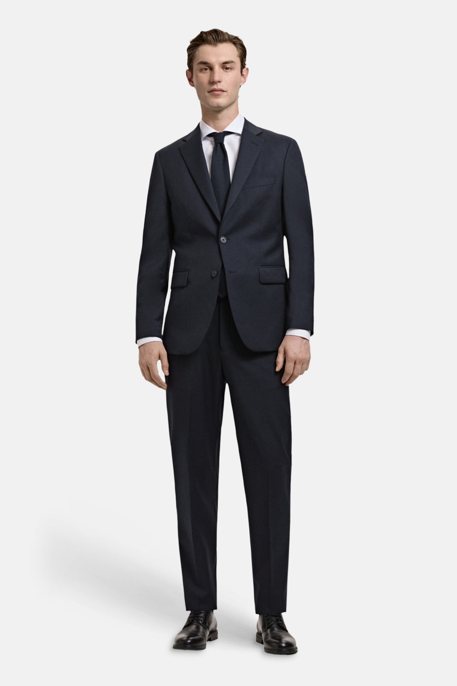 Chemise blanche à manches longues de MANGO, portée sous un costume bleu foncé avec cravate et chaussures noires.