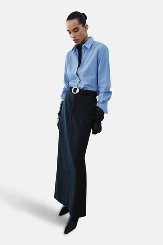Chemise bleue rayée à manches longues de MANGO, portée avec un gilet noir, une longue jupe, une ceinture et des gants.