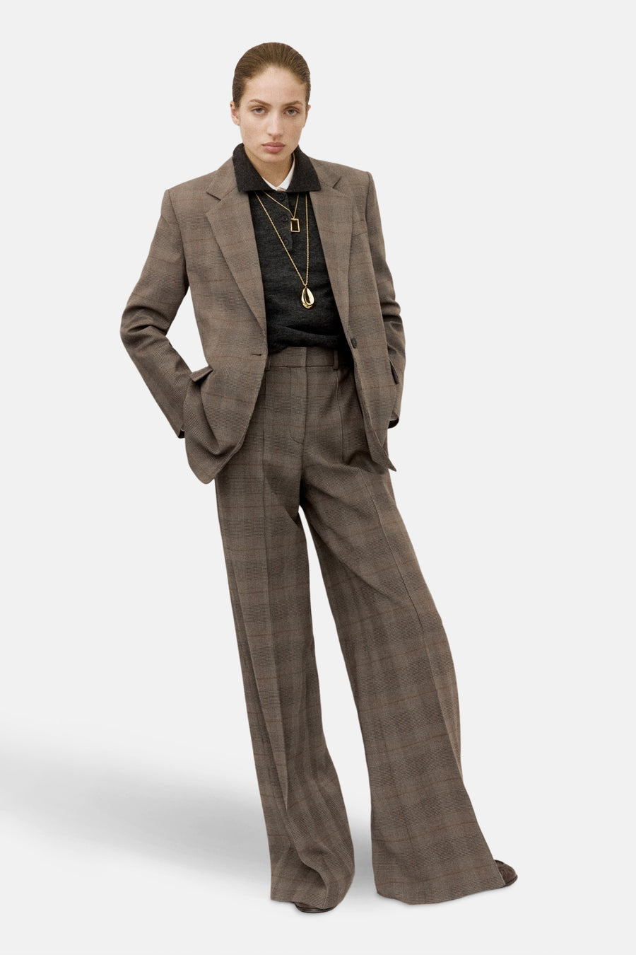 Blazer marron de MANGO, avec un motif à carreaux et porté avec une chemise sombre et des chaînes d'or.