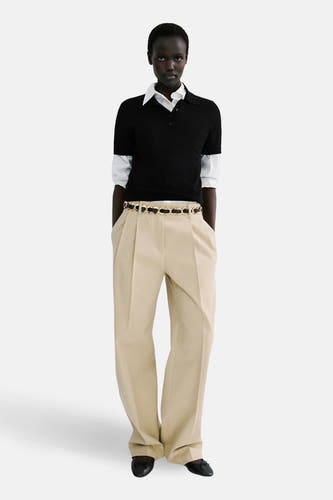 Polo noire à manches courtes de MANGO, portée sur une chemise blanche, avec une ceinture dorée sur un pantalon beige.
