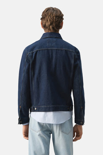 Donkerblauwe jeansjas van MANGO, achterkant gezien, gedragen met een gestreept overhemd en lichte jeans.
