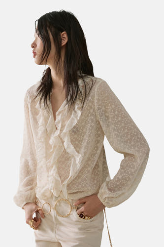 Chemisier écru à manches longues de MANGO avec motif floral brodé et col à volants, combiné avec un jean et une ceinture dorée.