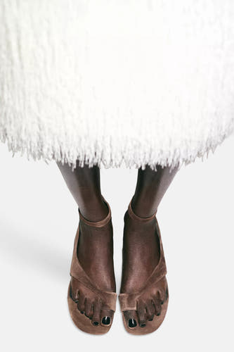 Sandales en daim marron de MANGO avec anneau d'orteil et bride à la cheville, visibles sur des pieds avec vernis à ongles noir sous une robe blanche à volants.