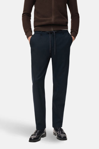 Man met bruin vest en donkerblauwe broek met trekkoord, staand.