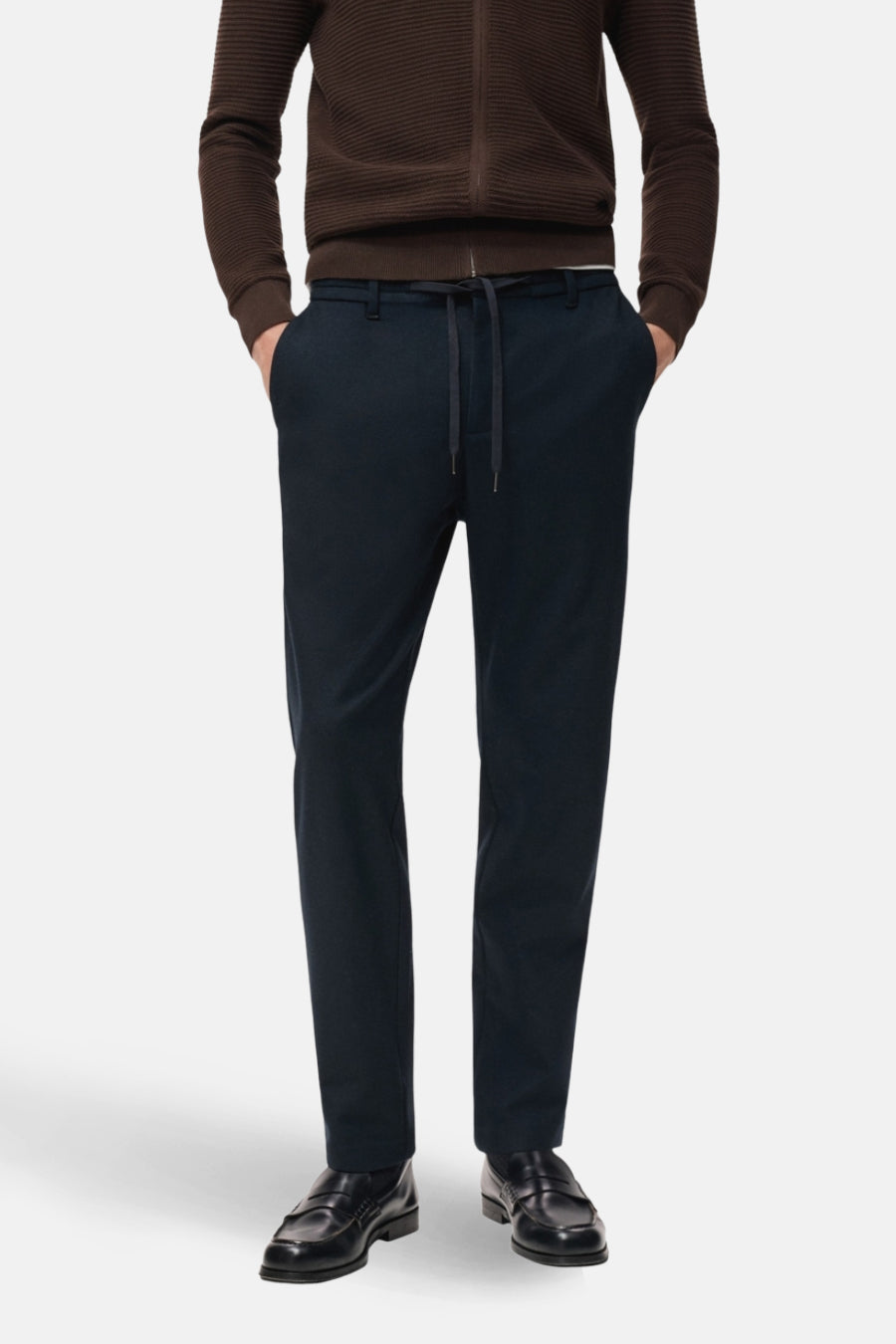 Man met bruin vest en donkerblauwe broek met trekkoord, staand.