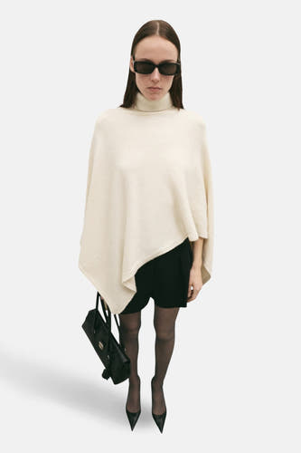 Poncho écru de MANGO, avec une coupe décontractée, porté sur une robe noire avec un string et des escarpins.
