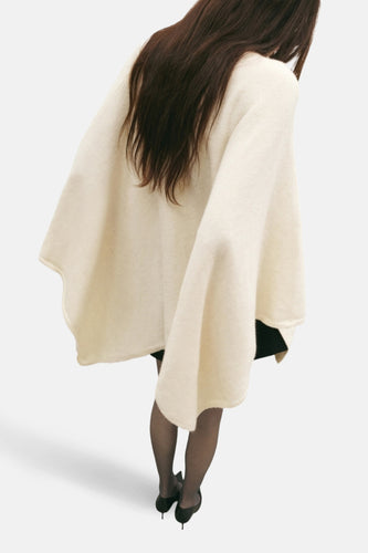 Vrouw met bruin haar draagt crèmekleurige poncho, zwarte panty en schoenen met hak.