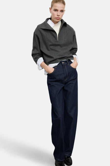 Vrouw in grijze trui, wit overhemd, donkerblauwe jeans, handen in zakken.