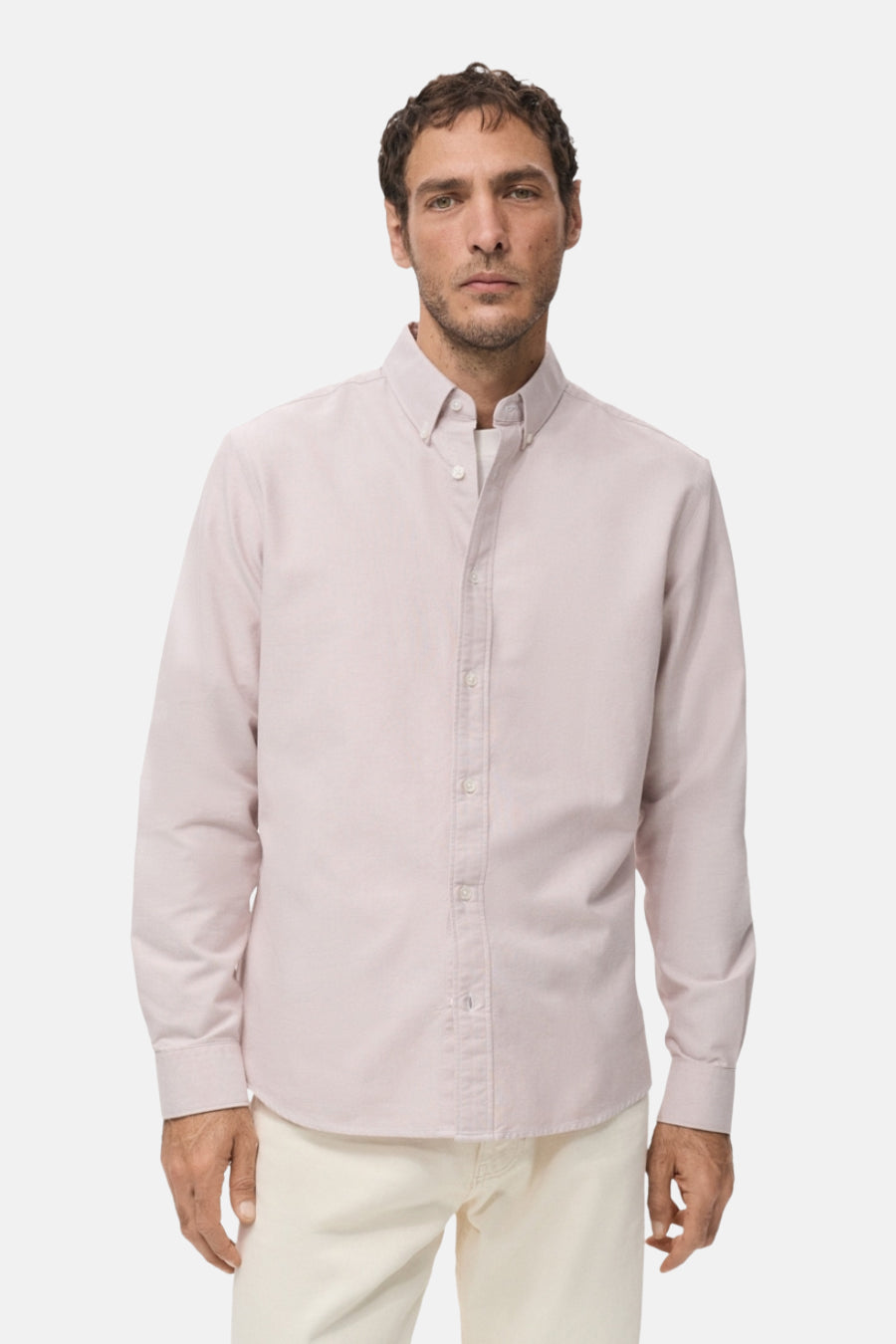 Man in roze overhemd en witte broek, staand.
