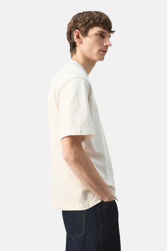 T-shirt écru à manches courtes de MANGO, vue de profil avec mannequin en jean foncé et mains dans les poches.