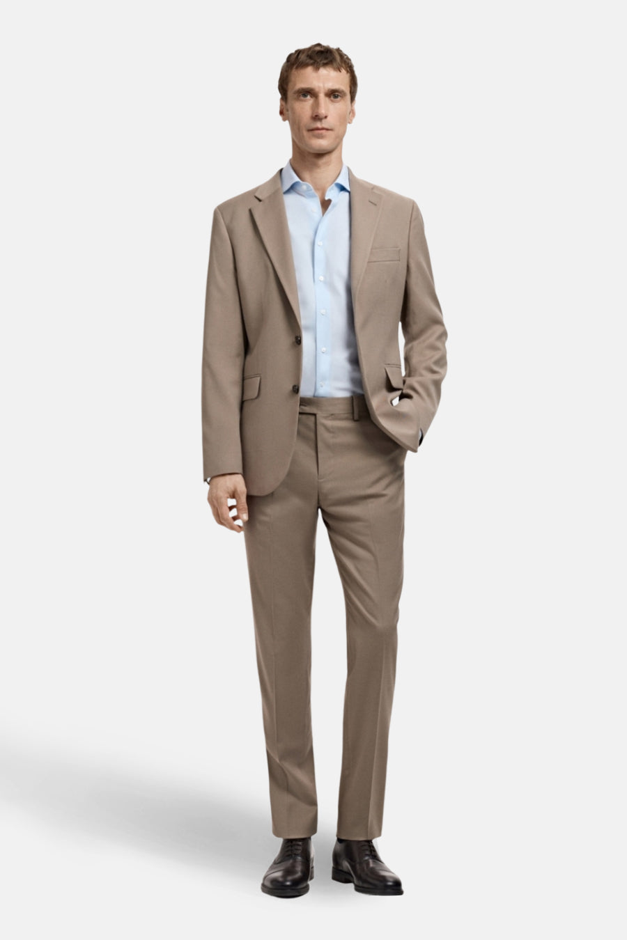 Man in bruin pak met blauw overhemd staand tegen witte achtergrond.