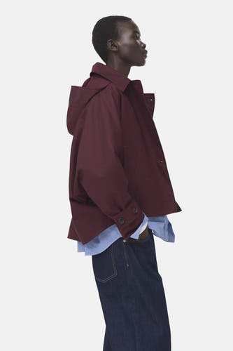 Manteau bordeaux de MANGO, avec capuche, porté sur une chemise rayée et un jean foncé.
