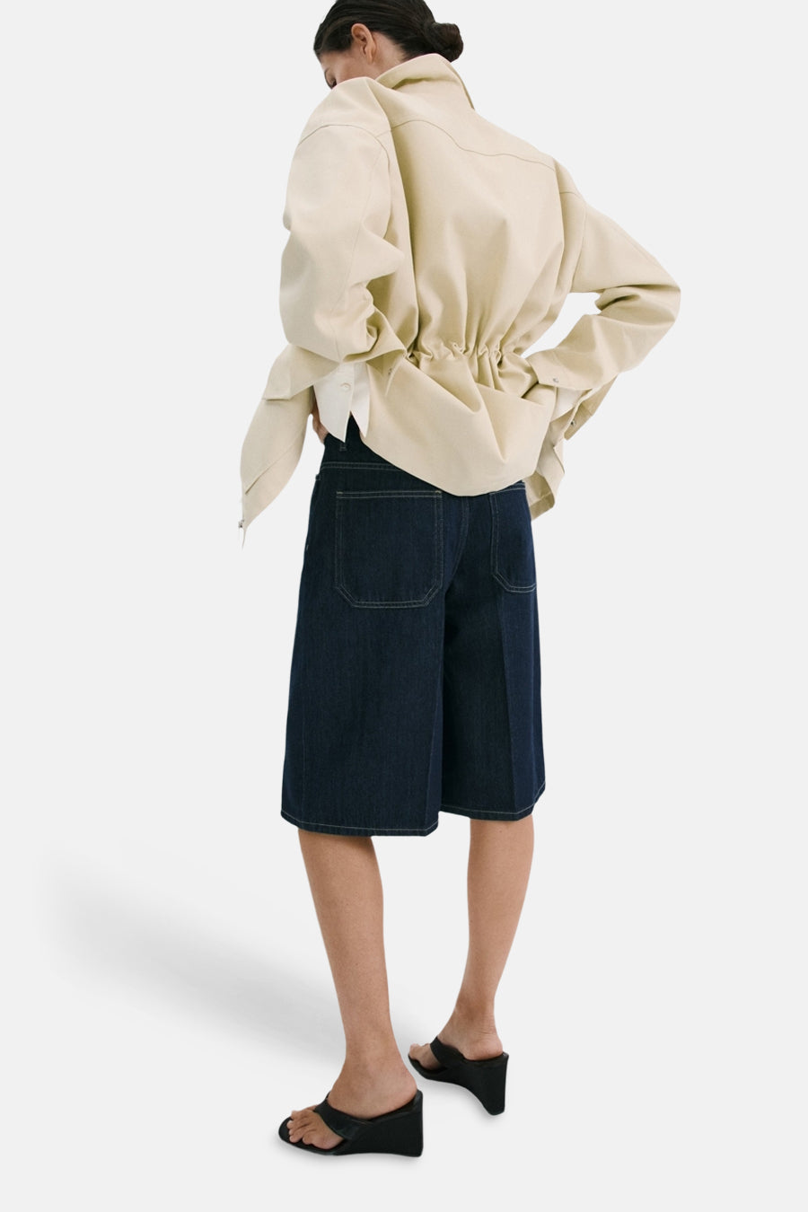 Donkerblauwe denim bermuda van MANGO, gezien van achteren, gecombineerd met een crèmekleurig jasje en zwarte sandalen.
