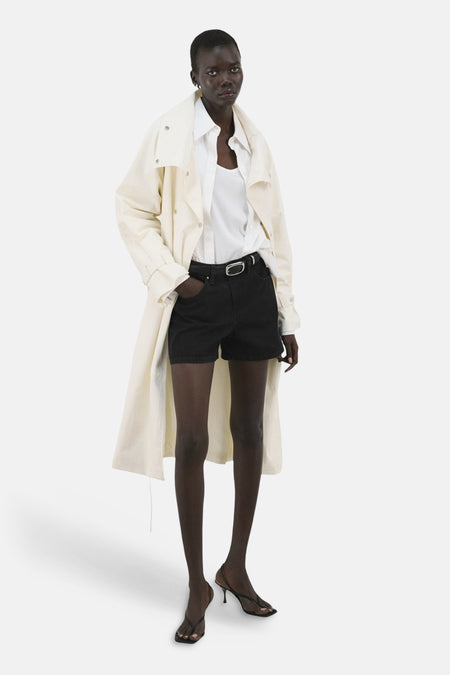 Bermuda short noire de MANGO, avec une boucle argentée et portée avec une blouse blanche et un trench-coat.