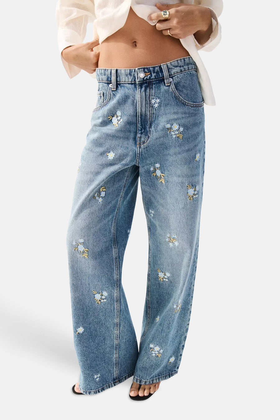 Jean droit en denim bleu clair de MANGO, avec broderies florales, porté à l'arrière par une femme en chemise blanche.