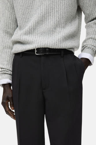 Pantalon noir de MANGO, avec plis et une fine ceine noire, associé à un pull gris.