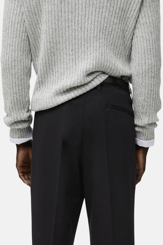 Pantalon noir de MANGO, avec une poche arrière, porté avec un pull gris et une chemise blanche.
