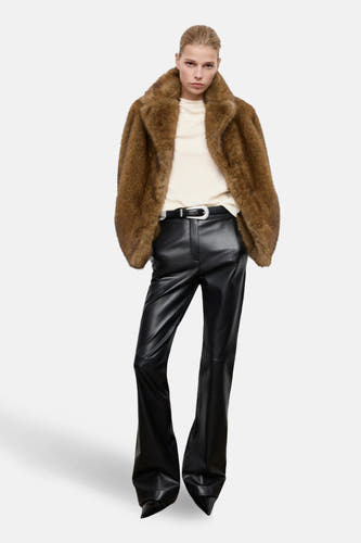 Blouse écru à longues manches de MANGO, portée sous un manteau mouton imité brun et une jupe en cuir noir avec ceinture.