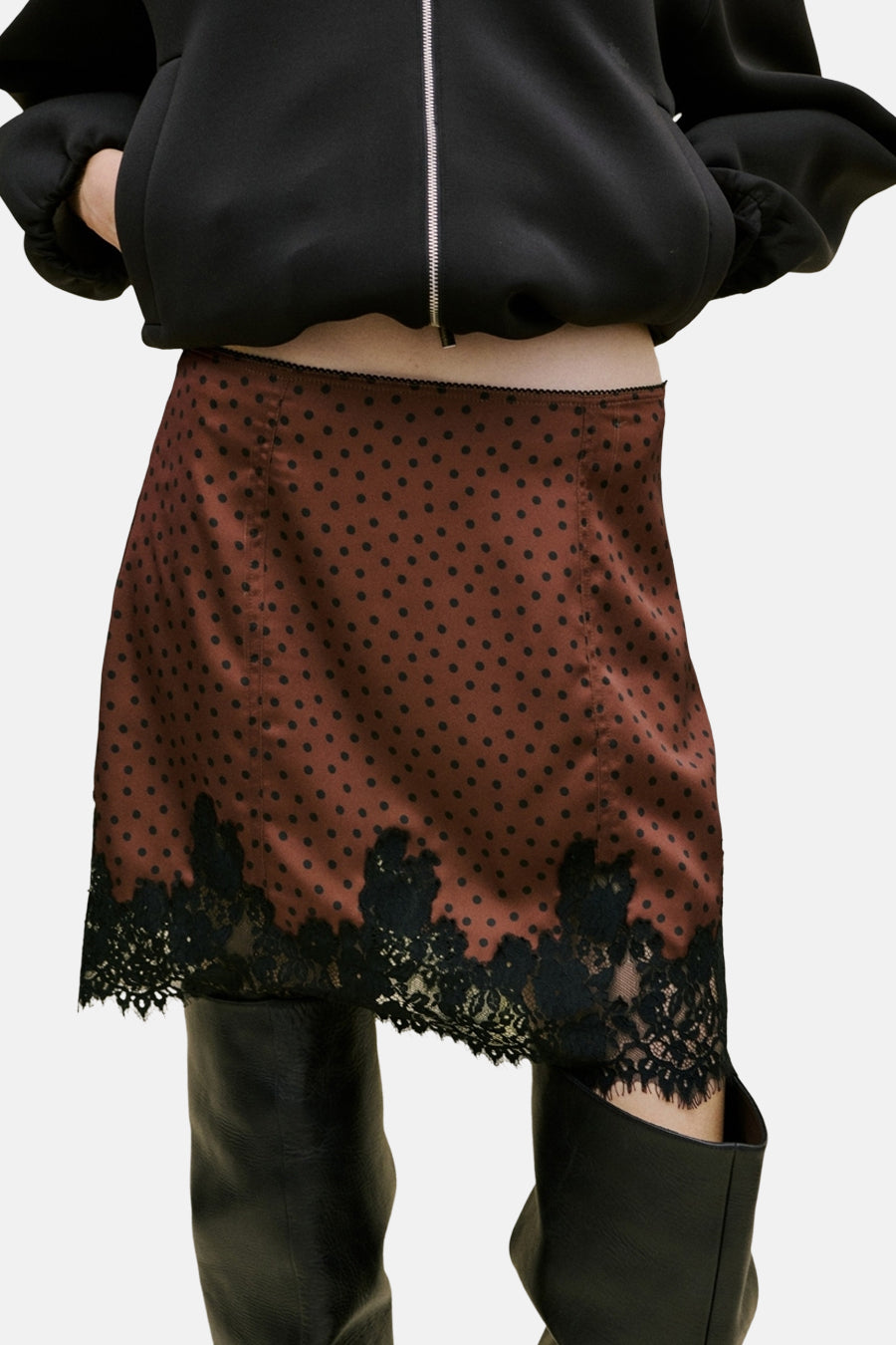 Jupe courte marron de MANGO, à motif à pois et bordure en dentelle noire, combinée avec des bottes noires et une écharpe blanche.